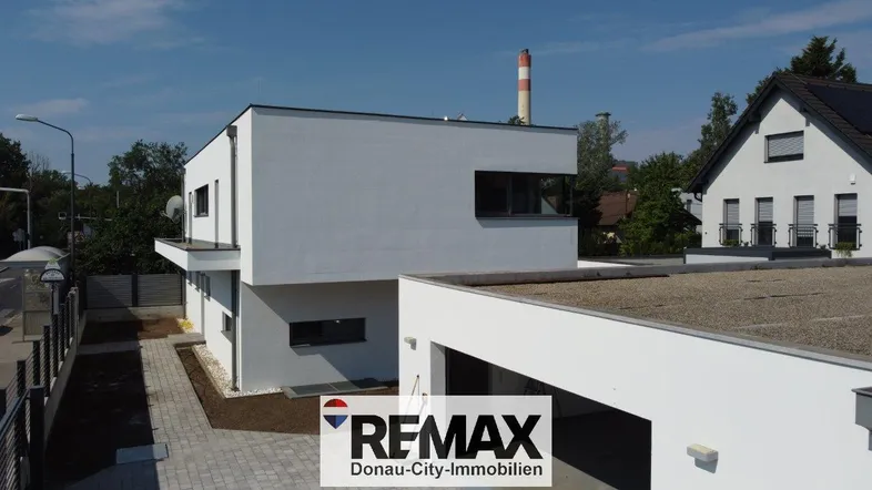 Modernes Einfamilienhaus mit Flachdach und Garage, umgeben von Grünflächen und einem Schornstein im Hintergrund.