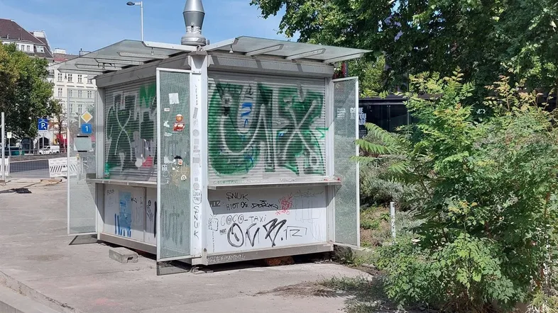 Außenansicht eines kleinen Kiosks mit Graffiti und Rollläden, umgeben von Bäumen und einer Straße.