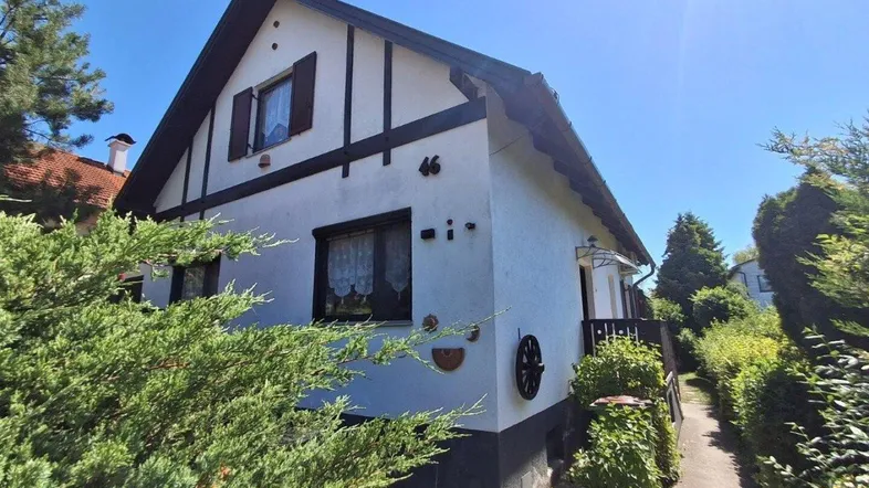 Weißes Haus mit dunklen Holzbalken und Satteldach, umgeben von grünen Büschen unter blauem Himmel.