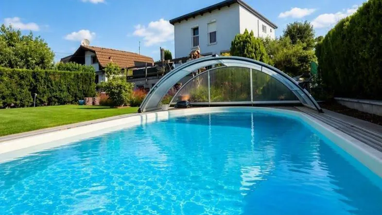 Großer Außenpool mit Schiebeüberdachung und gepflegtem Gartenbereich unter blauem Himmel.