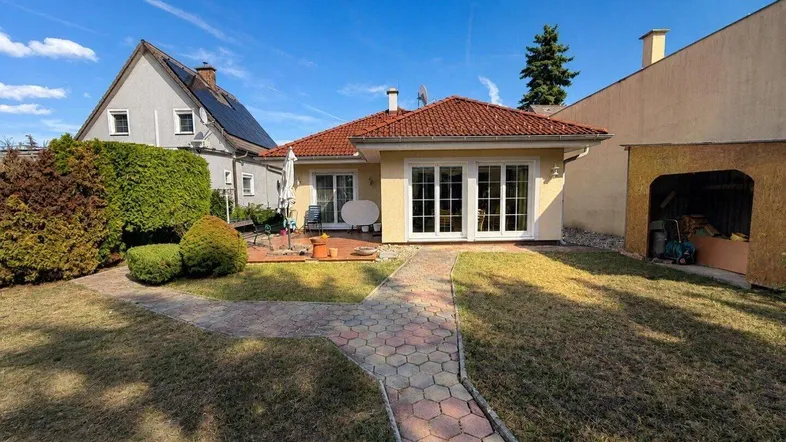 Blick auf das Haus von hinten mit Garten, Rasenfläche und gepflastertem Weg unter blauem Himmel.
