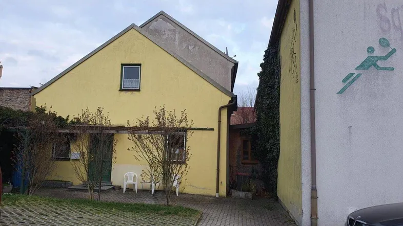 Gelbes Gebäude mit schrägem Dach und gepflastertem Vorplatz, daneben ein weiteres Gebäude.