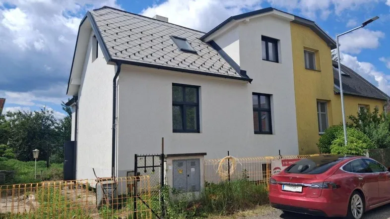 Weißes Haus mit dunklem Dach und rotem Auto davor, umgeben von Baustelle.