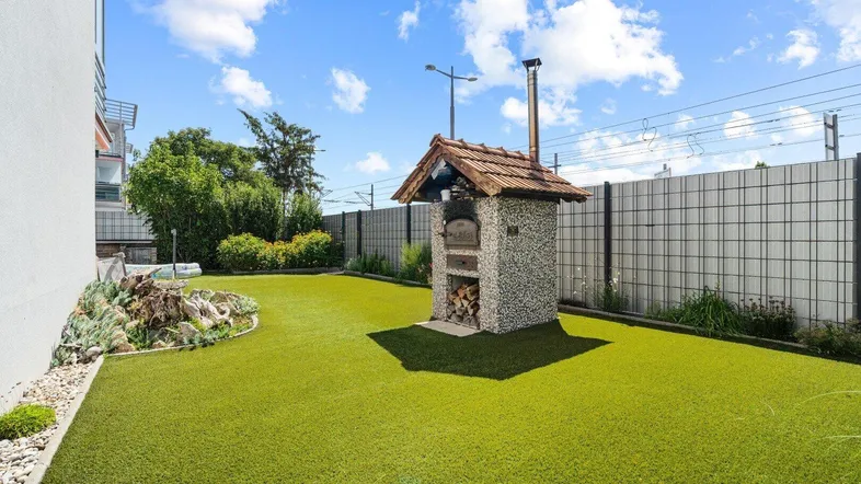 Großer Gartenbereich mit Kunstrasen, einem Steinofen und einer gepflegten Hecke.