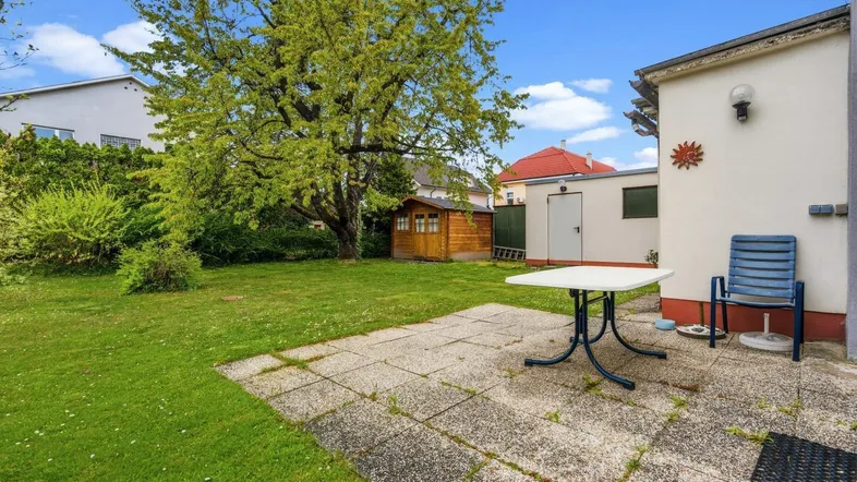 Großer Garten mit gepflasterter Terrasse, Rasenfläche und einem kleinen Holzschuppen im Hintergrund.