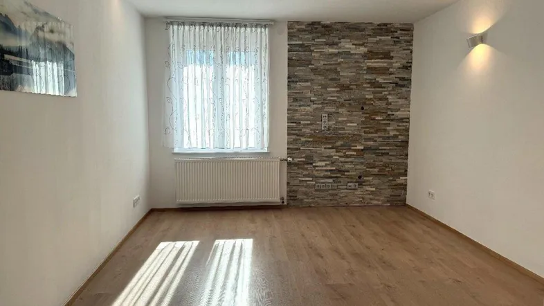 Helles Zimmer mit Holzboden, einer Steinwand und großem Fenster, ideal als Wohn- oder Schlafbereich.