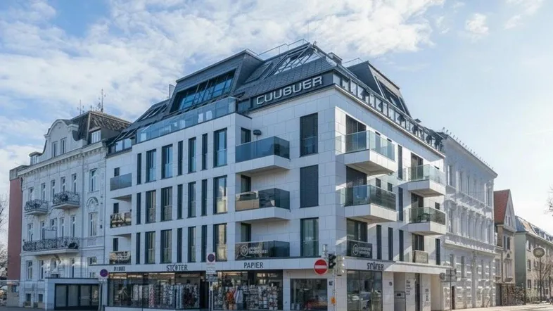 Modernes Wohngebäude mit Glasbalkonen und Geschäften im Erdgeschoss unter blauem Himmel.