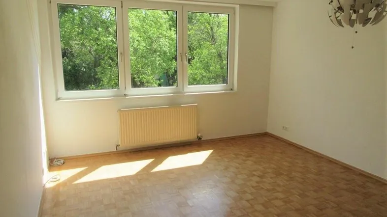 Helles Zimmer mit Parkettboden, Heizkörper und großem Fenster mit Blick ins Grüne.