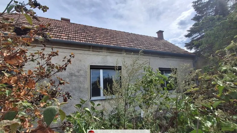Altes Haus mit Ziegeldach, umgeben von üppiger Vegetation und Bäumen.