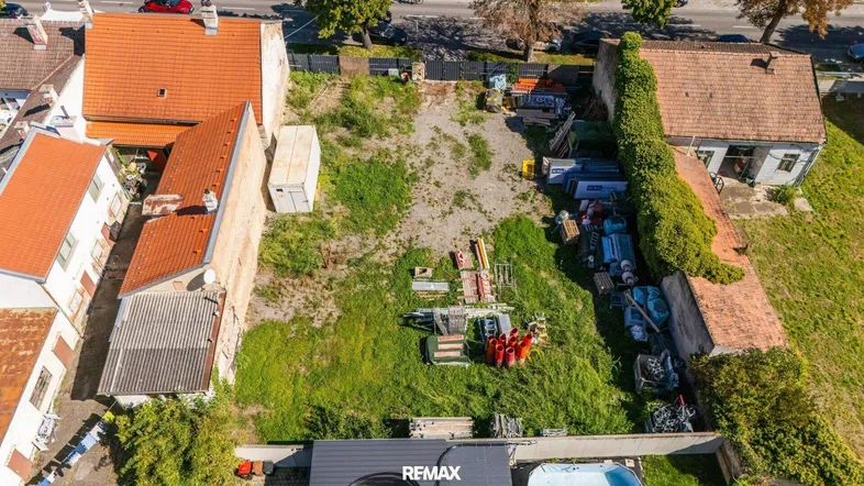 Luftaufnahme eines unbebauten Grundstücks mit umliegenden Gebäuden und Baumaterialien unter blauem Himmel.