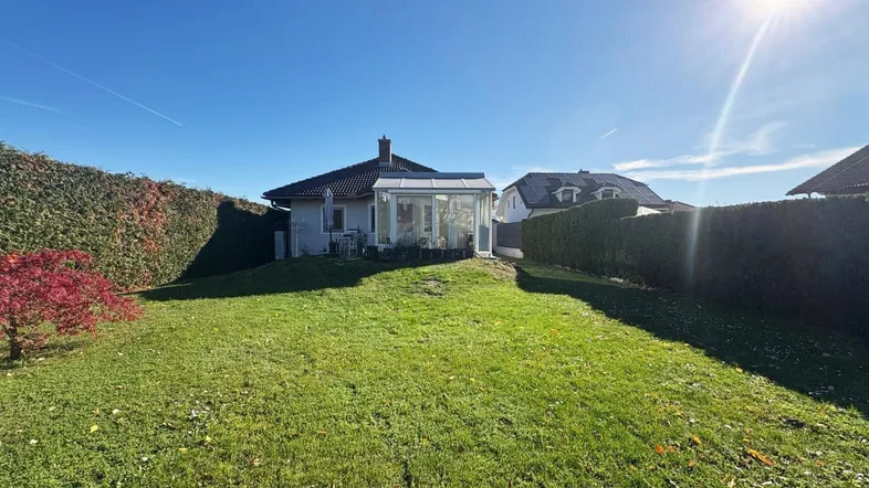 Sonniges Grundstück mit gepflegtem Rasen, Hecke und einem Wintergarten am Haus.
