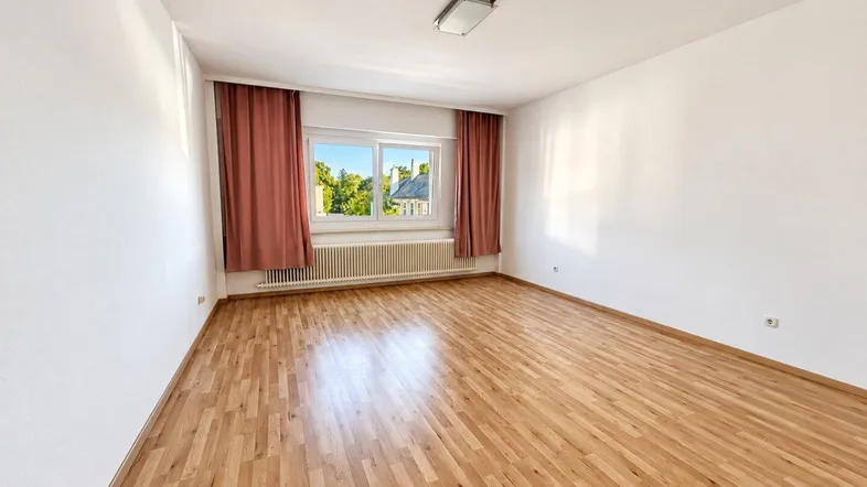 Helles Zimmer mit Parkettboden, weißen Wänden und einem Fenster mit roten Vorhängen.