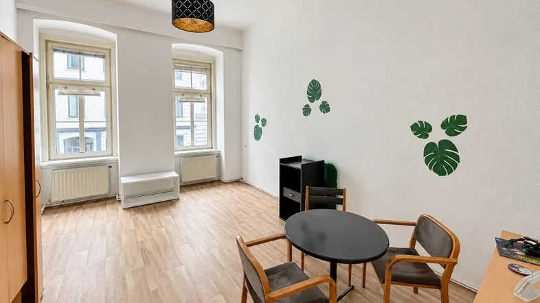 Helles Zimmer mit Laminatboden, hohen Fenstern und einfacher Einrichtung.