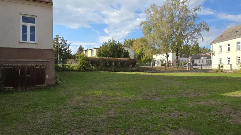 Große Rasenfläche mit angrenzendem Gebäude und weiteren Häusern im Hintergrund unter blauem Himmel.