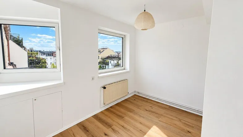 Helles Zimmer mit Holzboden, weißen Wänden und zwei Fenstern mit Blick auf die Stadt.