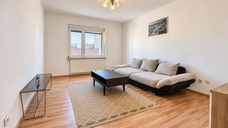 Helles Wohnzimmer mit beigem Sofa, Holzboden und einem Fenster, das Tageslicht hereinlässt.