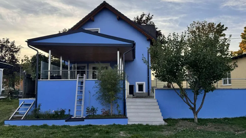 Blau gestrichenes Einfamilienhaus mit überdachter Terrasse und Gartenbereich unter einem bewölkten Himmel.