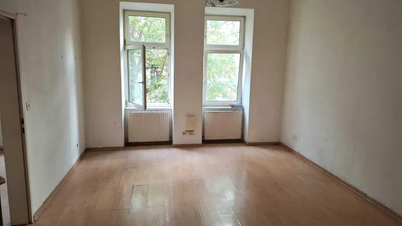 Leeres Wohnzimmer mit Holzboden, zwei großen Fenstern und Heizkörpern, in originalem Zustand.