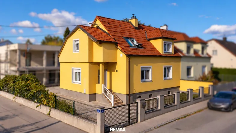 Gelbes Einfamilienhaus mit rotem Ziegeldach und gepflegtem Vorgarten an einer Wohnstraße.