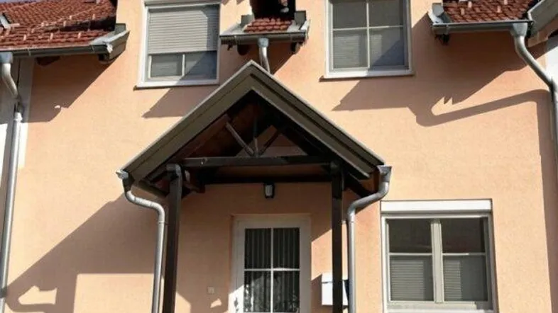 Vorderansicht eines Einfamilienhauses mit hellbeiger Fassade, weißen Fenstern und einem kleinen Vordach.