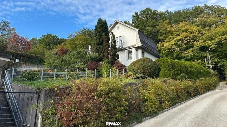 Weißes Haus mit Mansarddach, umgeben von üppiger Vegetation und Bäumen auf einem Hügel.