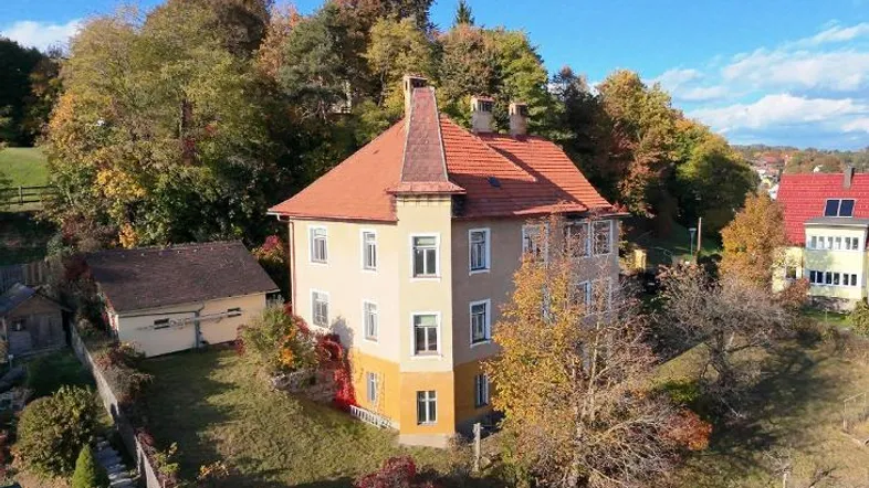 Historisches Gebäude mit markantem Turm und rotem Ziegeldach, umgeben von herbstlicher Vegetation.