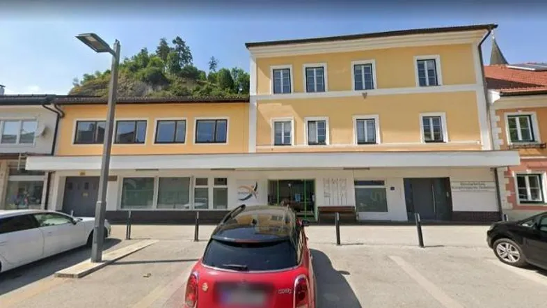 Gelbes Gebäude mit mehreren Etagen und Parkplätzen davor, umgeben von Bäumen und einem Hügel.