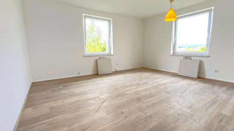 Helles Zimmer mit Laminatboden, weißen Wänden und Blick ins Grüne durch zwei Fenster.