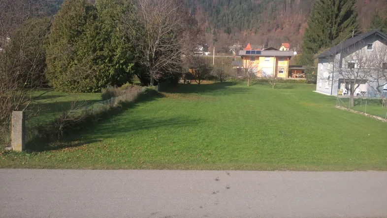 Grüne Wiese mit umliegenden Häusern und Bäumen, umgeben von einer bergigen Landschaft.