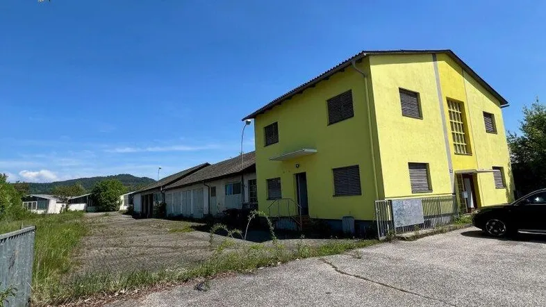 Gelbes Gebäude mit großem Vorplatz und weiteren Gebäuden im Hintergrund unter blauem Himmel.
