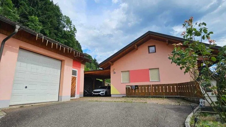 Rosa gestrichenes Haus mit Garage und Carport, umgeben von grüner Natur und Bäumen.