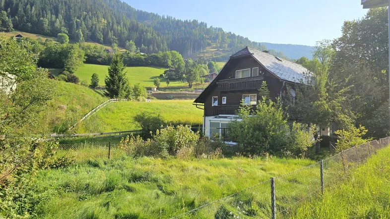 Traditionelles Holzhaus in ländlicher Umgebung mit grünen Wiesen und bewaldeten Bergen im Hintergrund.