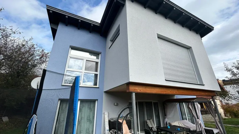 Modernes Haus mit blauer und grauer Fassade, einer möblierten Terrasse und einem Trampolin im Garten.
