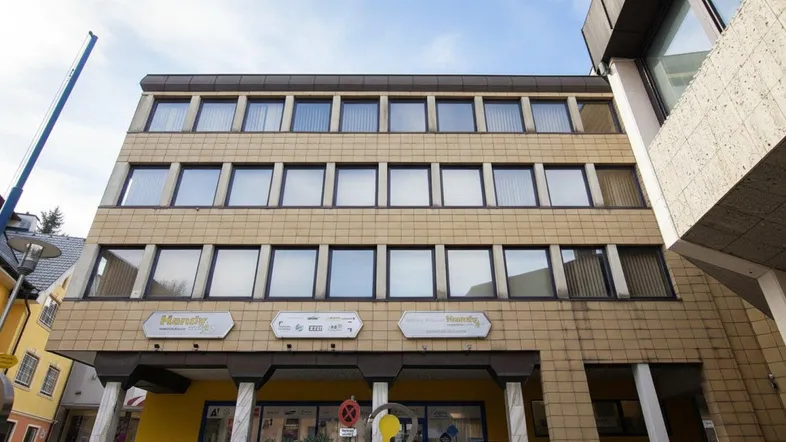 Mehrstöckiges Gebäude mit gelblicher Fassade und vielen Fenstern, im Erdgeschoss befinden sich Geschäfte.