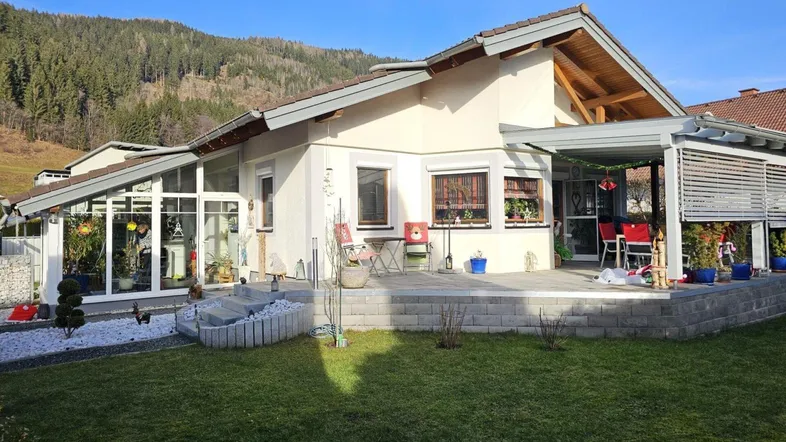 Modernes Haus mit großer Terrasse, Wintergarten und gepflegtem Garten vor bewaldetem Hügel.