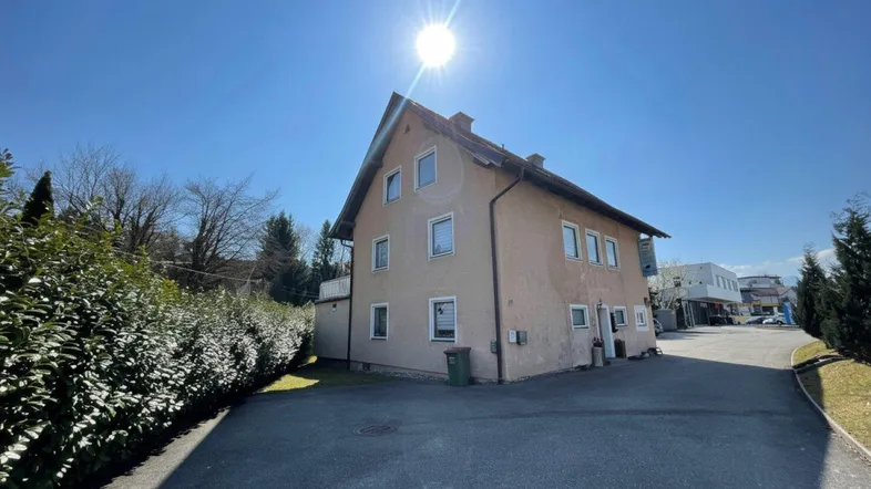 Gebäudeansicht von der Seite mit Zufahrt und gepflegter Hecke unter strahlendem Sonnenschein.