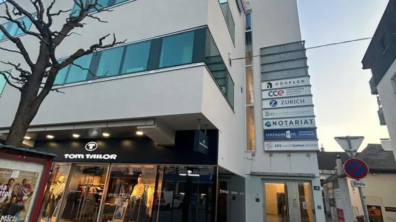 Modernes Geschäftsgebäude mit großen Fenstern und Ladenflächen im Erdgeschoss bei Dämmerung.
