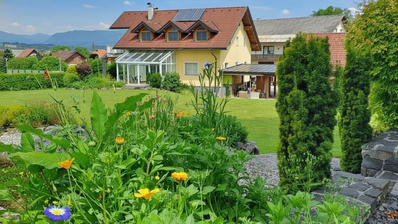 Gepflegter Garten mit blühenden Pflanzen, Rasenfläche und Blick auf das Haus mit Wintergarten.