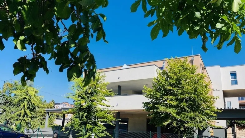 Modernes Wohngebäude mit Balkonen, umgeben von Bäumen und Grünflächen unter blauem Himmel.
