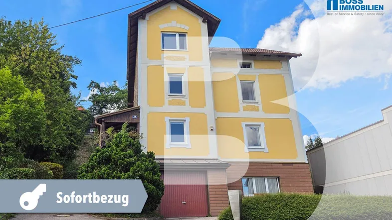 Gelbe Fassade eines Mehrfamilienhauses mit rotem Garagentor und grüner Bepflanzung.