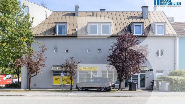Graues Gebäude mit Mansarddach und Geschäftsflächen im Erdgeschoss an einer Straße.