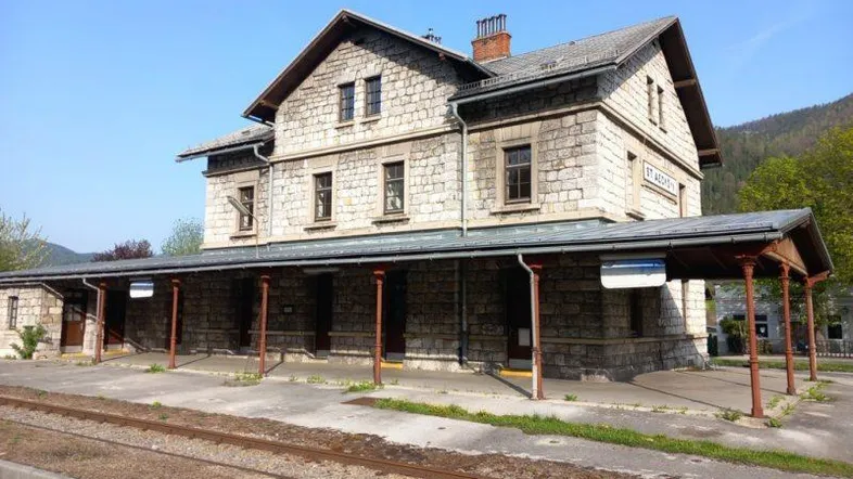 Historisches Steingebäude mit Bahnsteigüberdachung und Gleisen, umgeben von grüner Landschaft und Bergen.