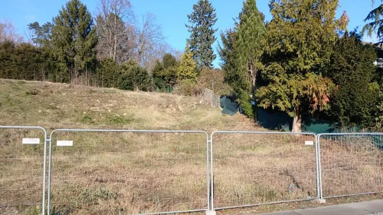 Leeres Grundstück mit Bauzaun und Baumbestand im Hintergrund unter blauem Himmel.