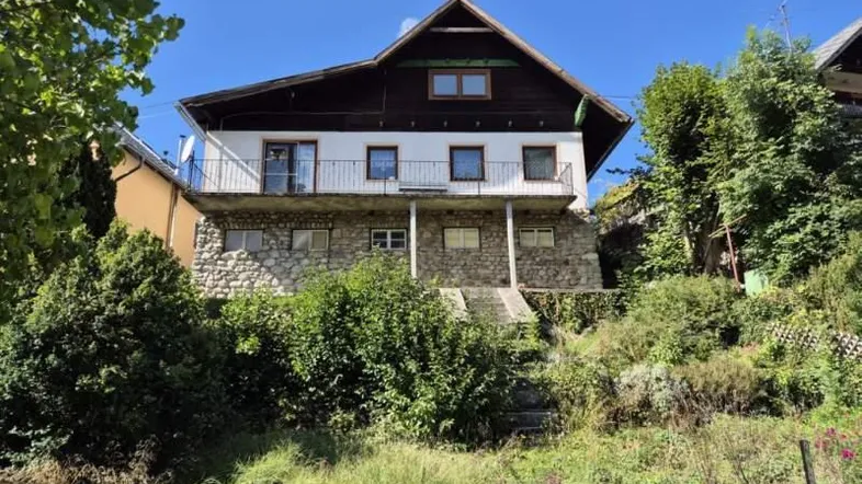 Hausfassade mit Steinsockel und Balkonen, umgeben von viel Grün unter blauem Himmel.