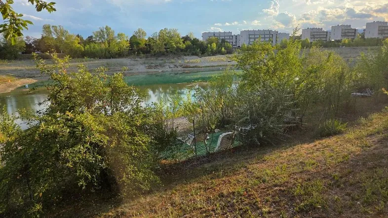 Blick auf einen See mit Ufervegetation und Wohngebäuden im Hintergrund unter einem bewölkten Himmel.