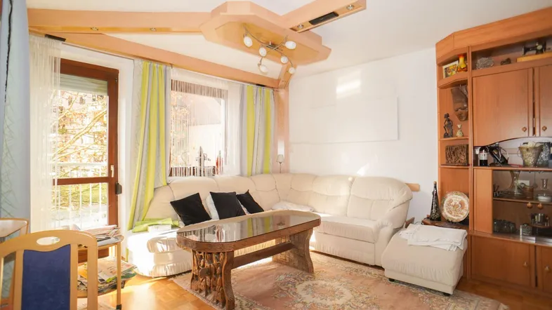 Helles Wohnzimmer mit beigem Sofa, Holzbalken an der Decke und Zugang zum Balkon.