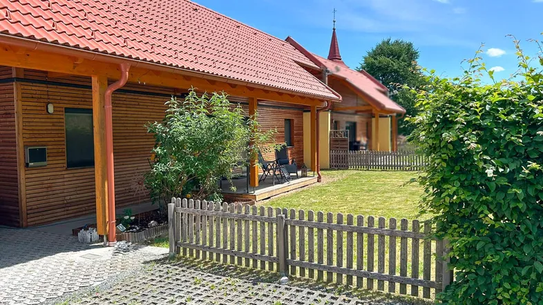 Holzverkleidetes Haus mit rotem Ziegeldach, umzäuntem Garten und gepflastertem Parkplatz.