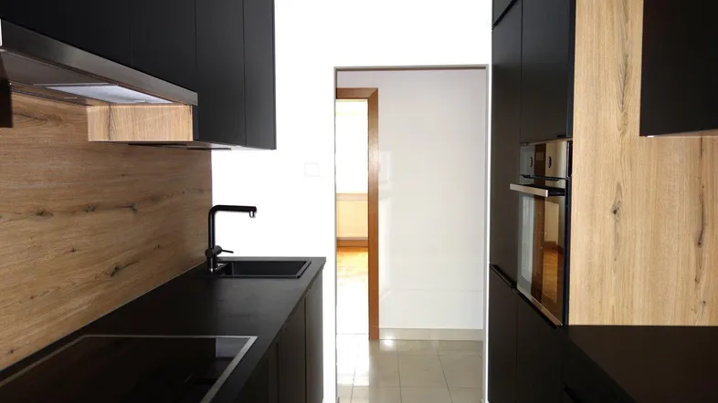 Moderne Küche mit schwarzen Schränken, Holzrückwand und integrierten Geräten, die zu einem weiteren Raum führt.