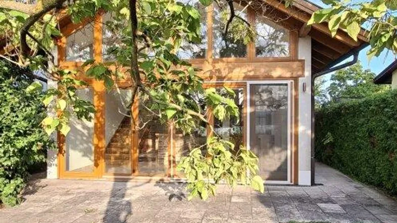 Außenansicht des Hauses mit großen Glasfronten, umgeben von Grün und einer gepflasterten Terrasse.