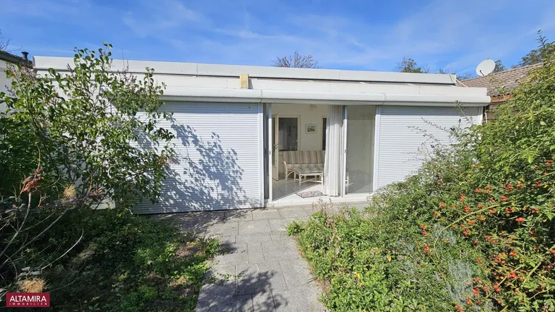 Kleines Gebäude mit Rollläden und Zugang zu einem gepflasterten Weg im grünen Gartenbereich.